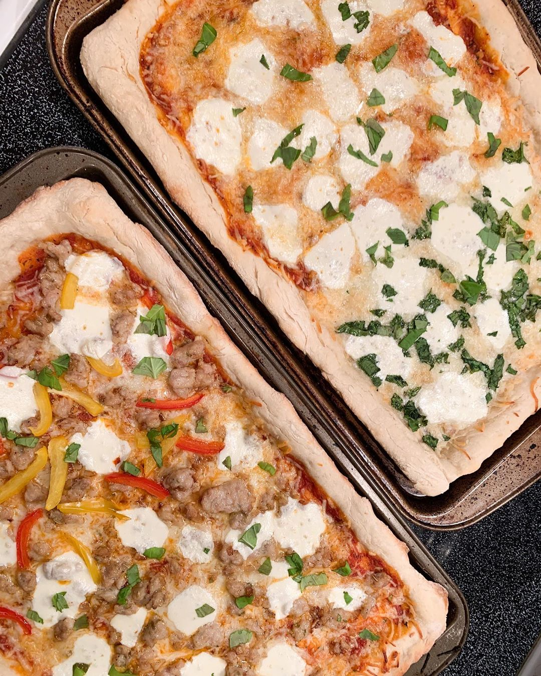 two rectangular pizzas topped with fresh mozzarella, herbs, veggies, and sausage.