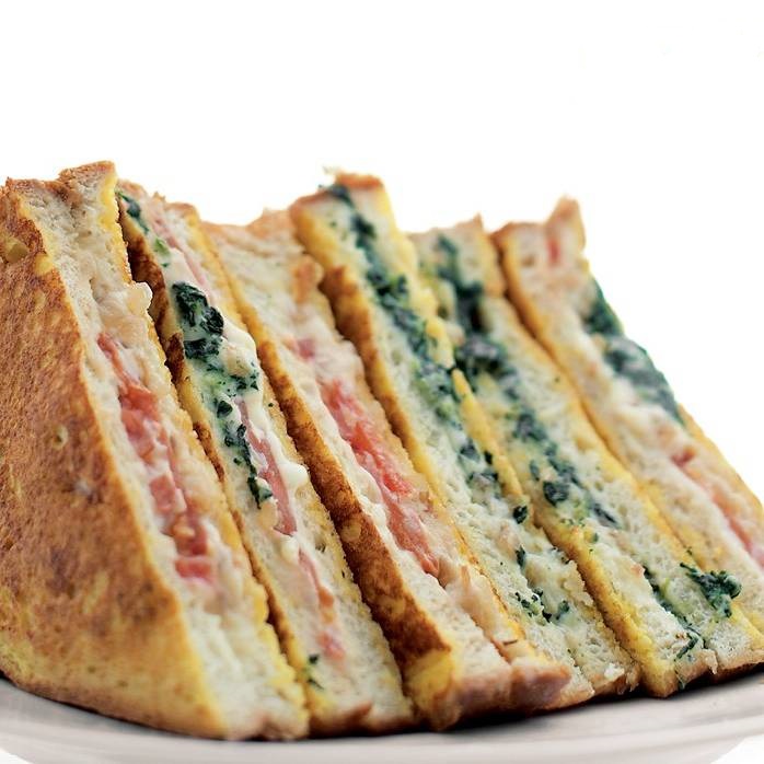 A photo of veggie and cheese stuffed french toast with golden brown bread sandwiched together. The interior of the sandwich is made up of melty cheese, tomatoes, and greens.