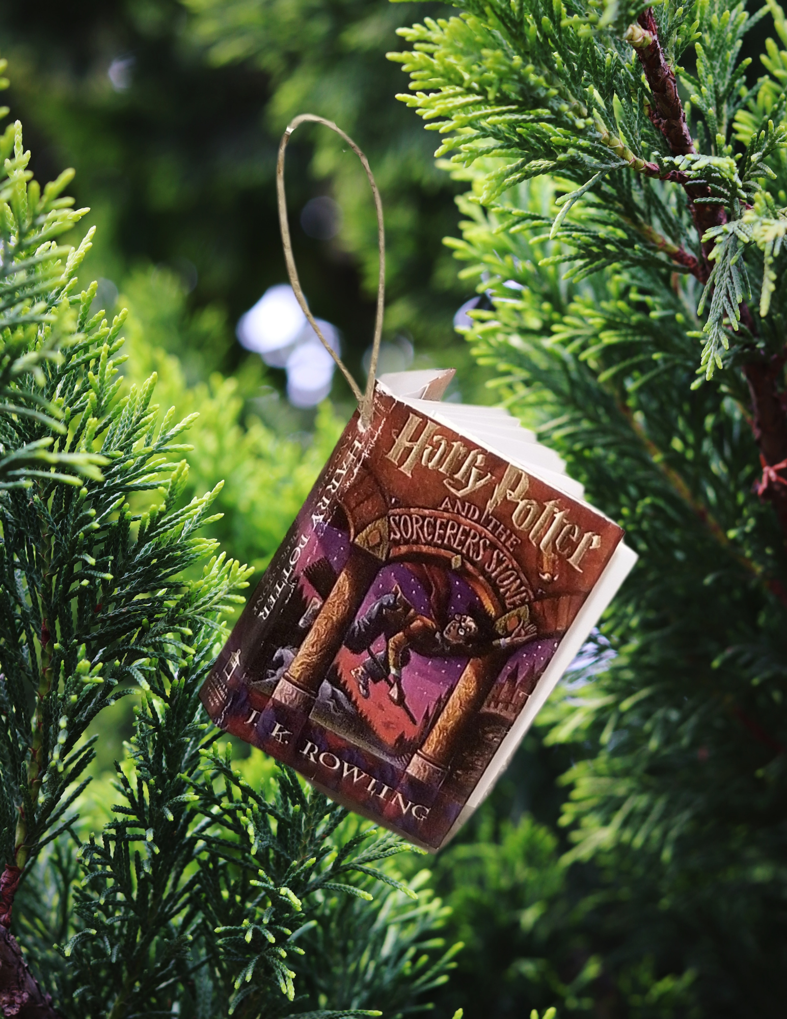 A miniature version of the book "Harry Potter and the Sorcerer's Stone" as an ornament in front of green evergreen foliage