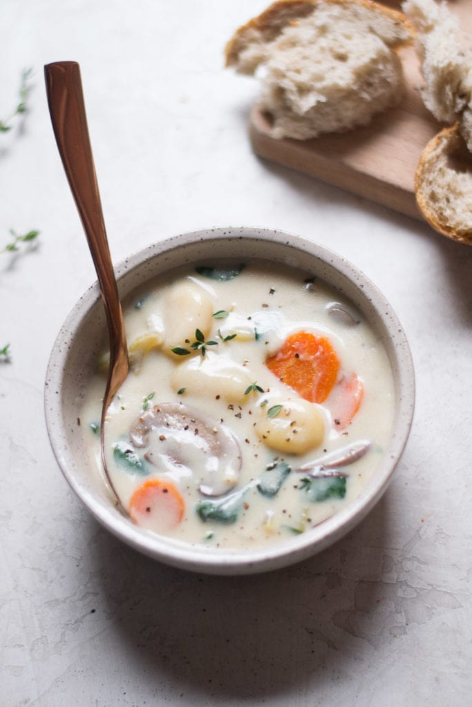 A bowl of creamy gnocchi and vegetable soup with crusty bread in the background