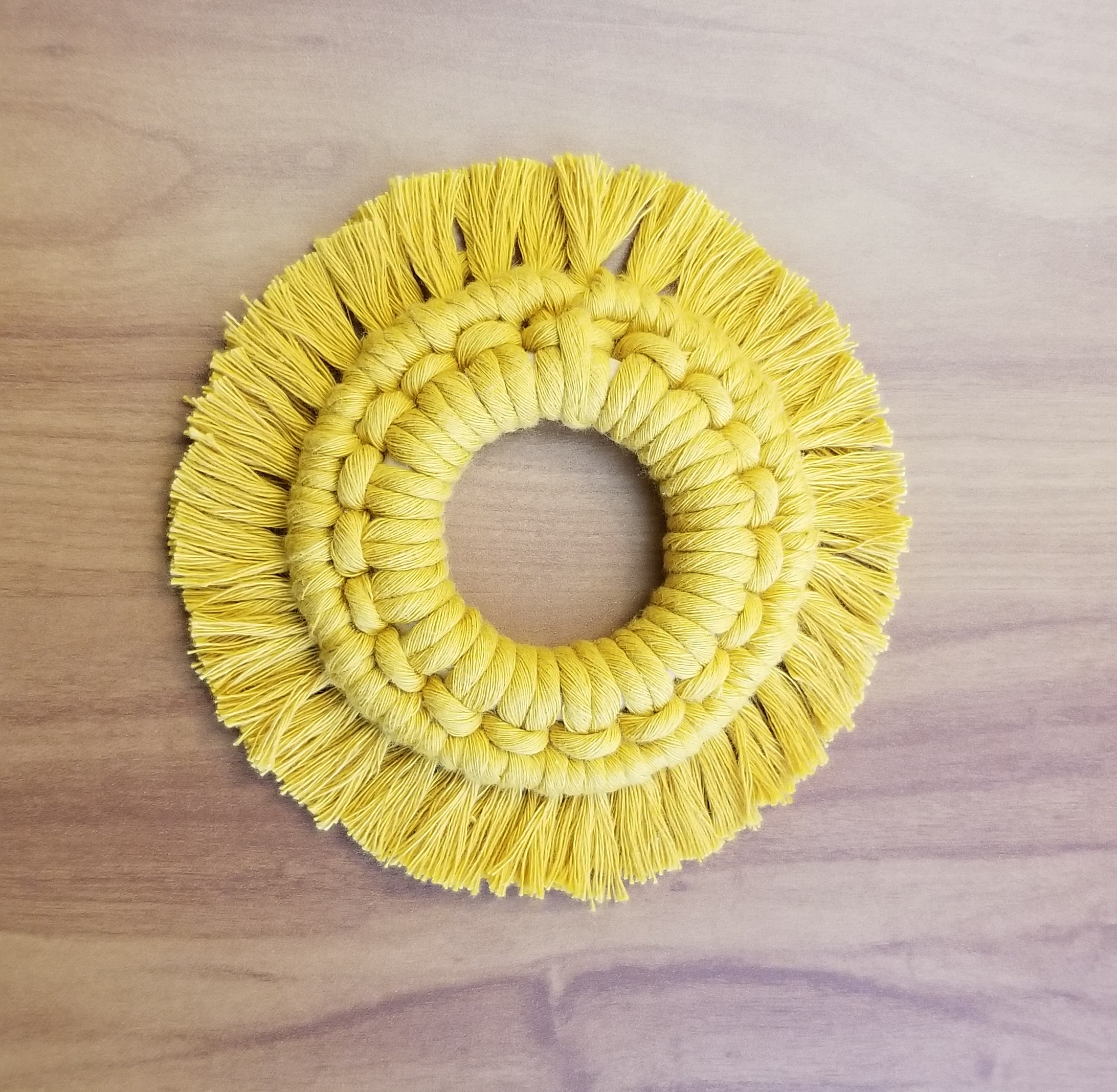 A yellow macrame sun wreath; it is a circle with an open center, and then repeating knots around a ring. Moving outward from the knots are fringe pieces, making the whole thing look like a sun.at the 