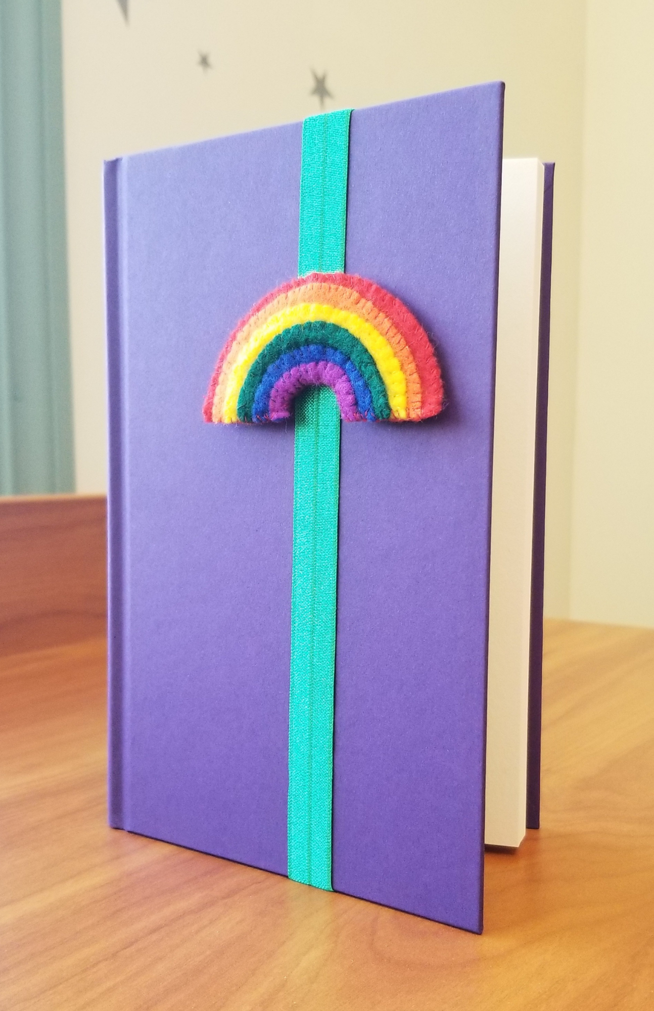A bookmark made of elastic wrapped around a book cover to hold the reader's place; there is a cartoony felt rainbow attached to the front of the bookmark