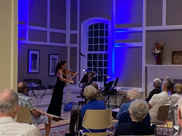 Alyce Cognetta Bertz on violin accompanied by her sister pianist Meg Cognetta Heaton. The backs of heads of audience members are visible.