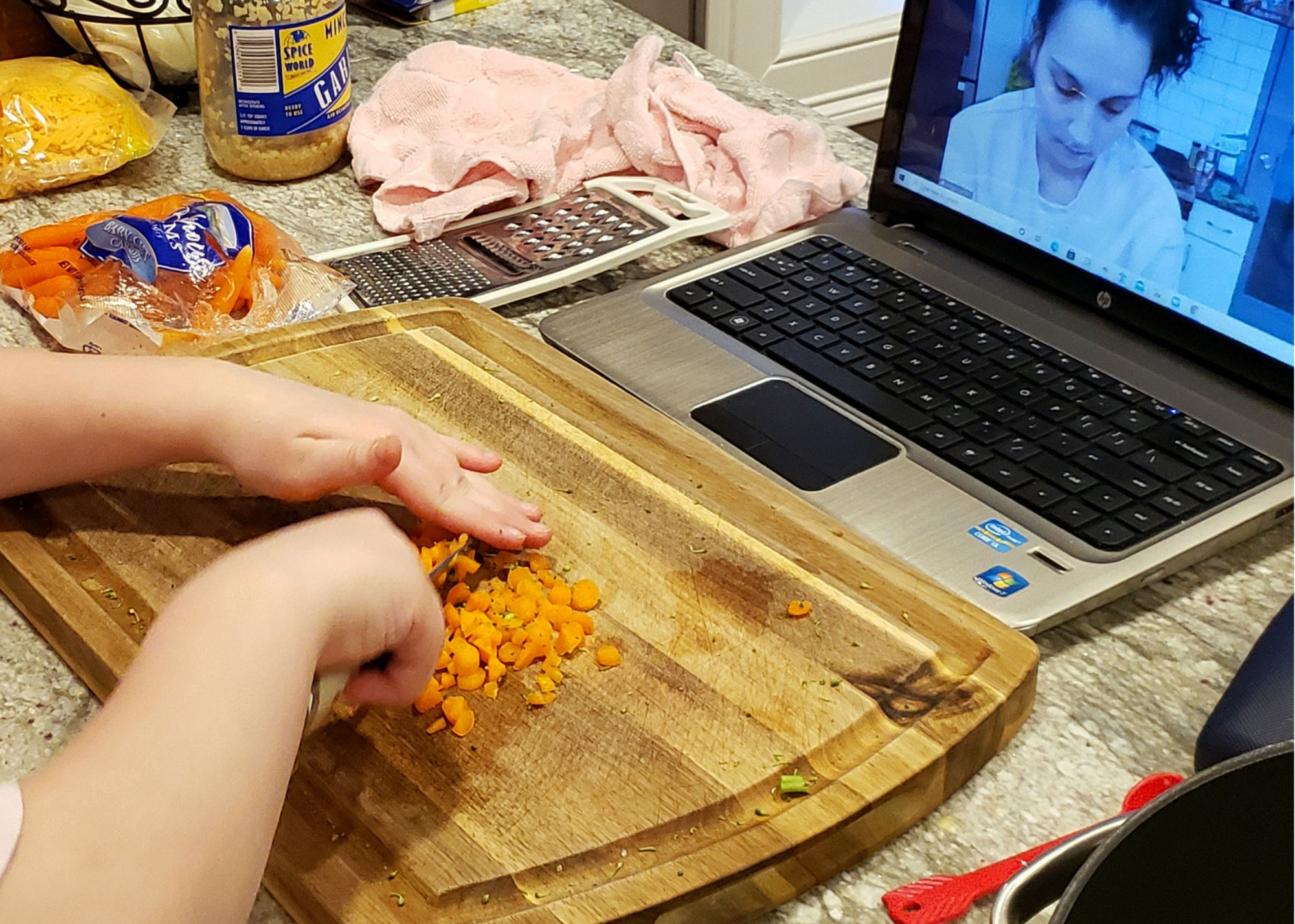 A child's hands are visible, cutting carrots on a wooded cutting board on a counter with cheese, garlic, more carrots, and a laptop with Food Explorers presenter Megan visible.