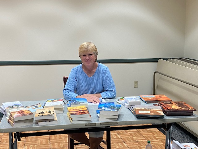 Author Nan Rossiter smiling and sitting behind a table with a display of her books ready to sign