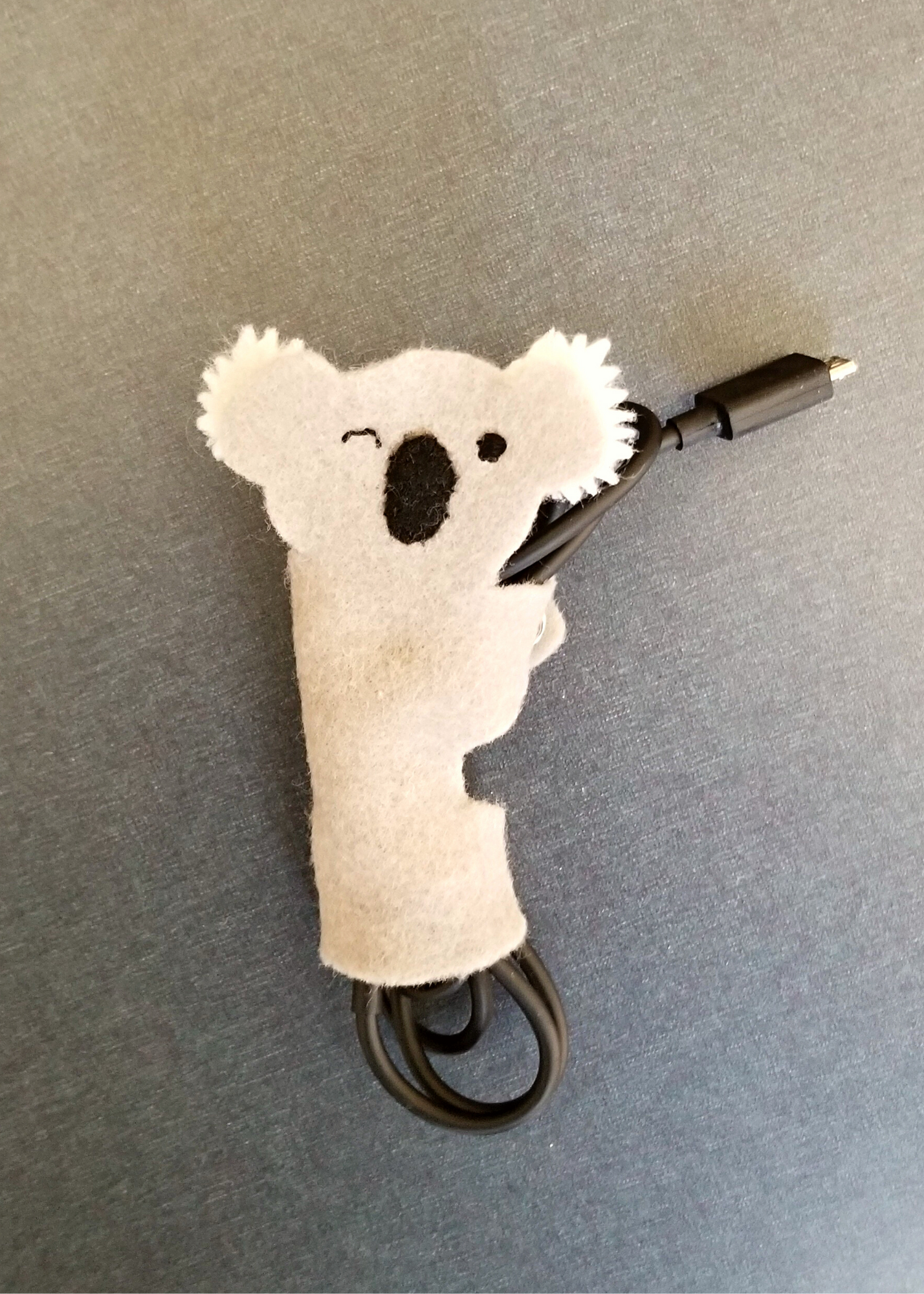 A gray felt koala with a winky face who is using its arms and legs to wind up a charging cord