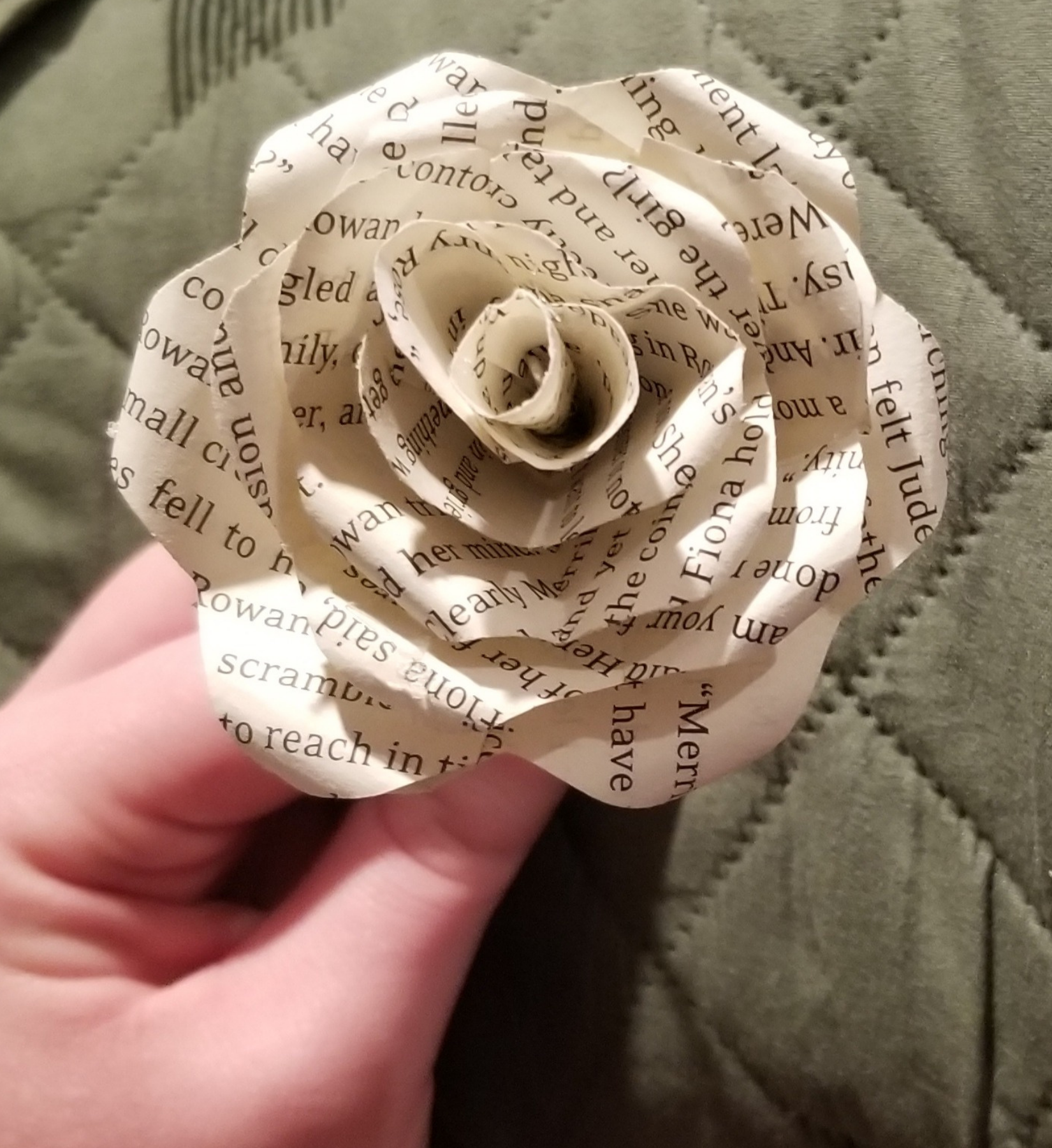 a rose made out of book pages held by a light skinned hand against a green background