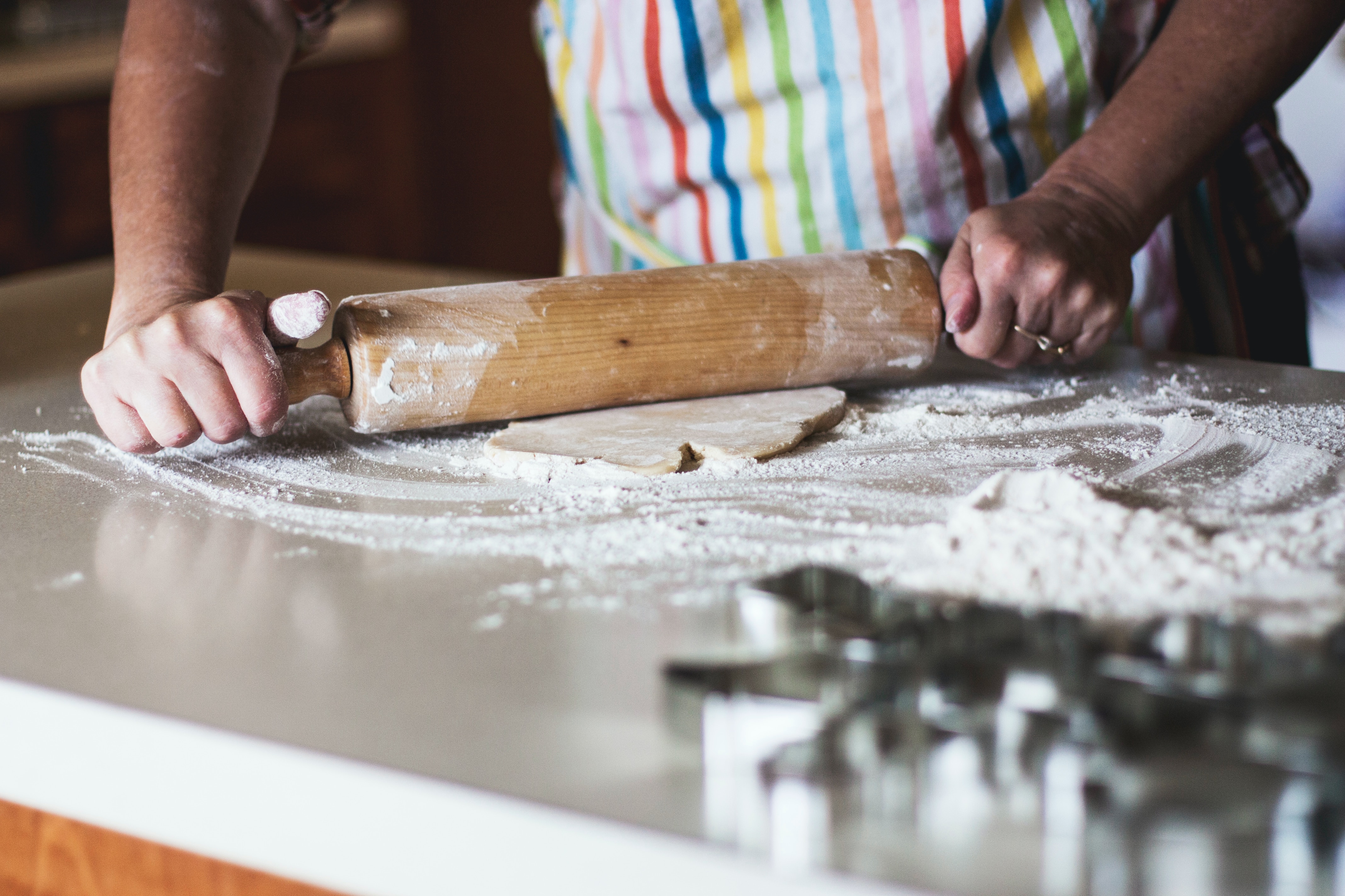 A person with light tan skin using a rolling pin on dough and flour; they are wearing a fun, rainbow striped apron.