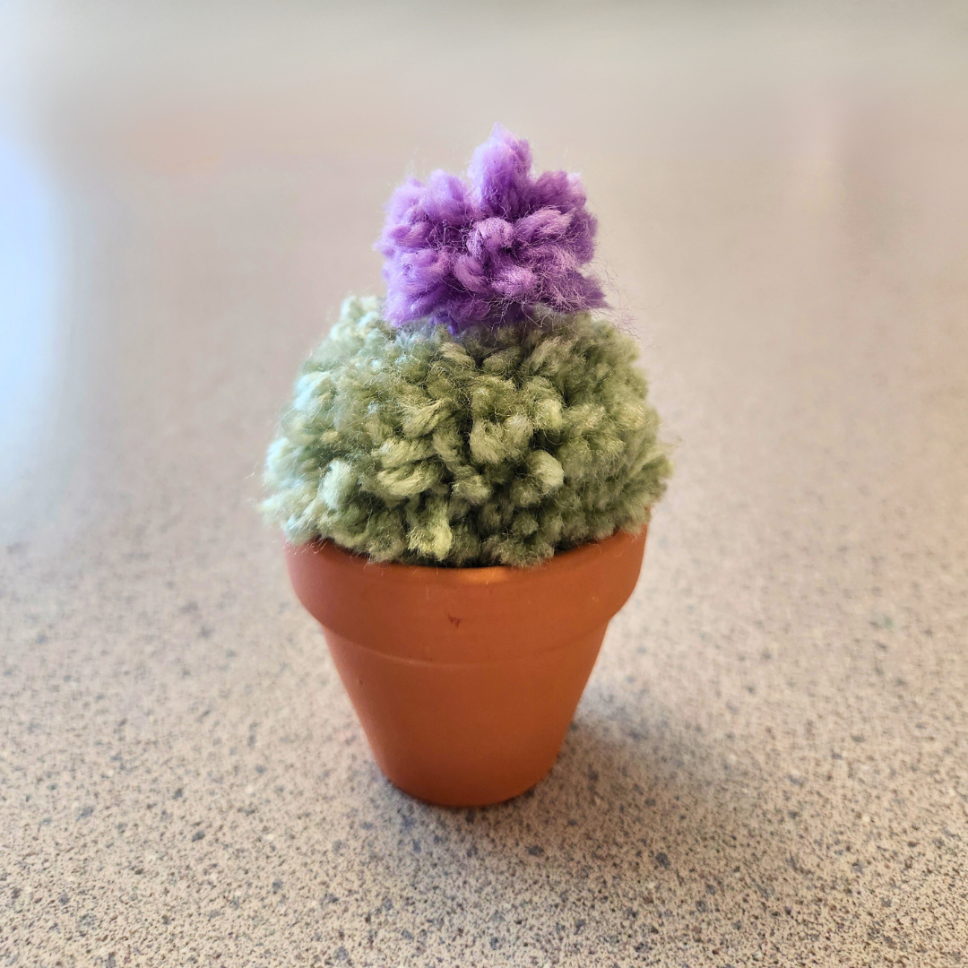 A sample of the craft, a small terracotta pot with a light green round pom pom with a purple flower on top; it resembles a small, round cactus.