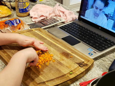 A child's hands are visible, cutting carrots on a wooded cutting board on a counter with cheese, garlic, more carrots, and a laptop with Food Explorers presenter Megan visible.